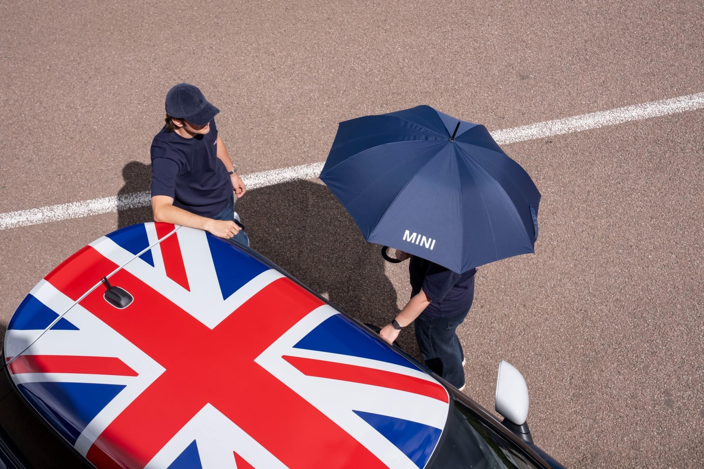 Vista superior do tejadilho Union Jack do MINI Cooper Oxford Edition, junto a duas pessoas, uma delas com um guarda-chuva MINI azul